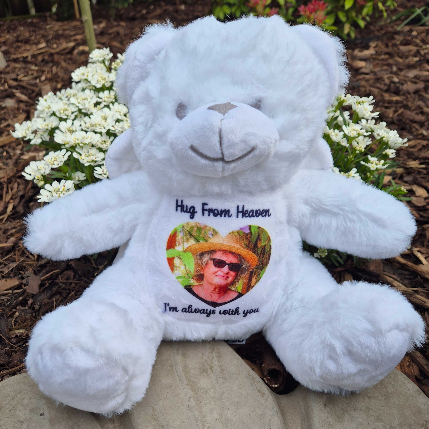 White angel teddy bear with a heart-shaped photo and text on a stone surface with flowers in the background CFHANG