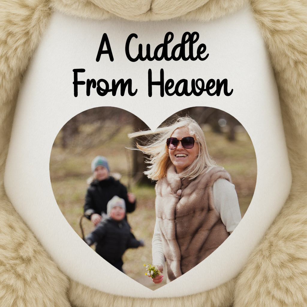 Teddy bear with printed Heart-shaped photo with a family picture and 'A Cuddle From Heaven' text, surrounded by fluffy beige fabric.
