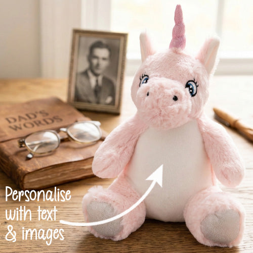 Pink plush unicorn toy on a wooden surface with a book and photo frame in the background.