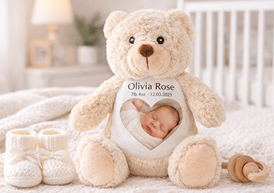 Beige teddy bear with a photo of a baby printed on it, placed on a soft surface with baby items around.