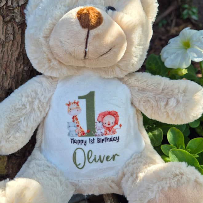 Brown teddy bear with a 'Happy 1st Birthday Oliver' shirt, surrounded by greenery and flowers.