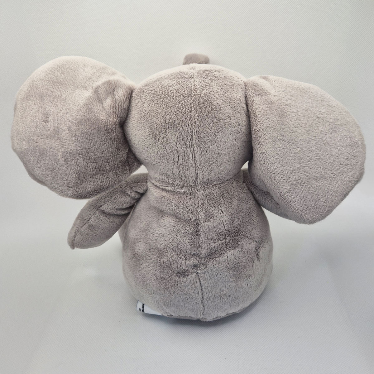 Back view of a grey plush elephant toy on a light grey background